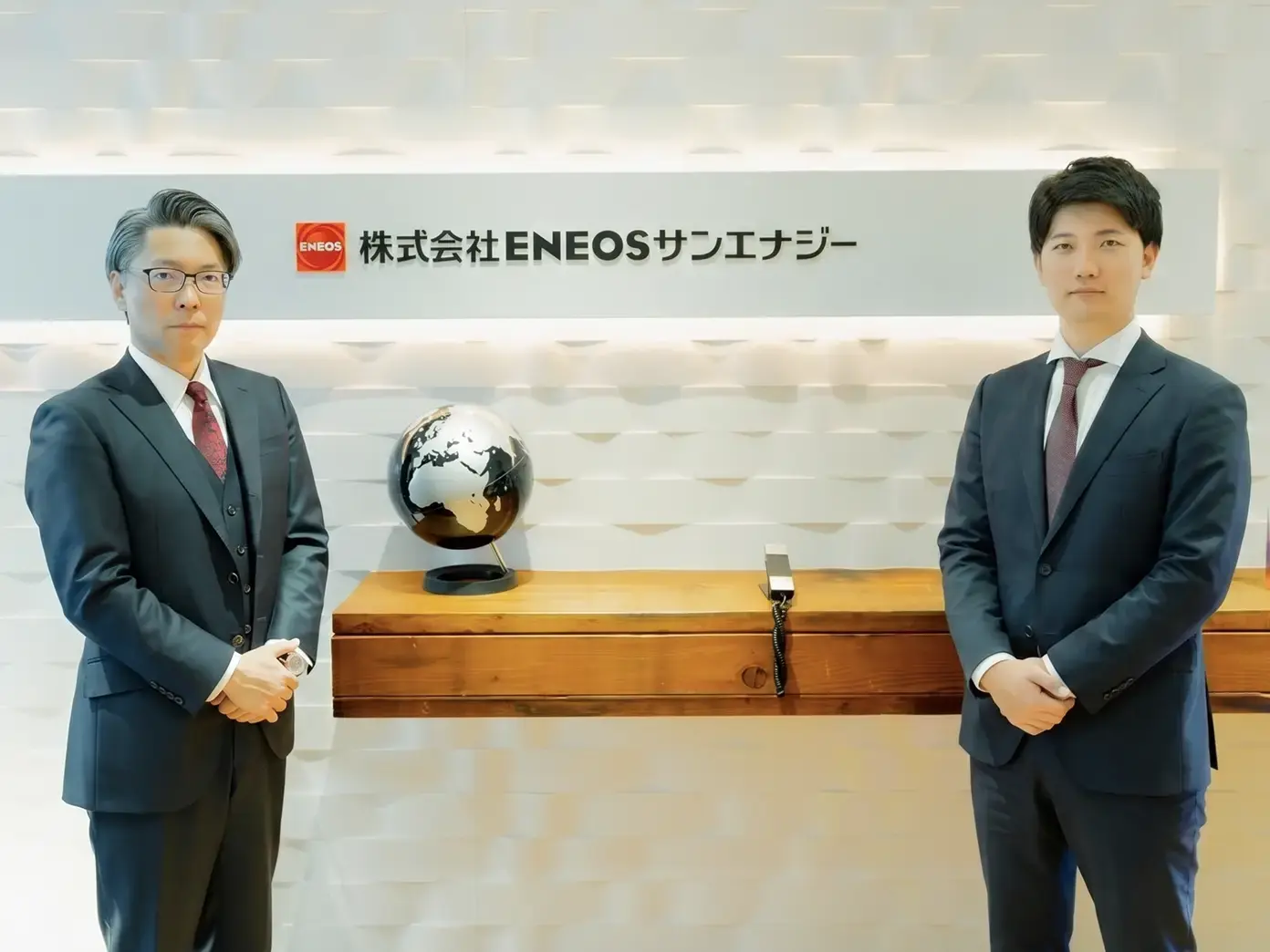 Two men in dark suits standing either side of a globe, beneath the ENEOS Sun Energy sign in a company reception.