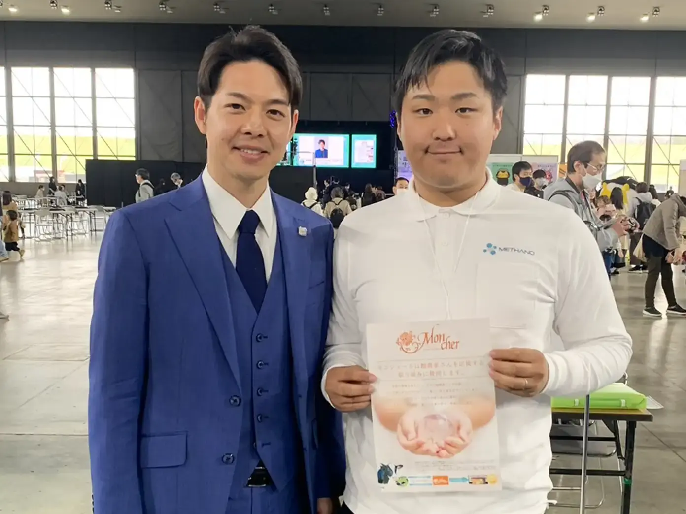 Two men side by side on the floor of an exhibition hall, one in a blue suit, the other in a white polo shirt holding a printed leaflet.
