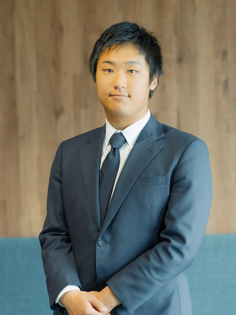 A man in a dark suit and tie, his hands clasped, standing in front of a wooden wall.